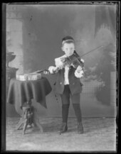 A "young boy playing the violin." Beside him is a table with likely a banjo on it.